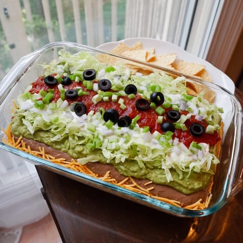 Chilled Seven-Layer Dip featuring guacamole, sour cream, and melted cheese topped with black olives and green onions.