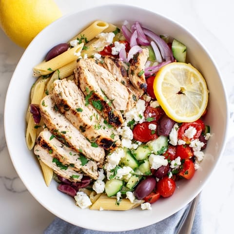 A close-up of a Greek Chicken Pasta Bowl featuring tender chicken, crisp cucumbers, briny Kalamata olives, and fresh parsley.  